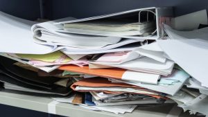A messy pile of papers and folders in disarray sit on a shelf, conveying chaos and clutter. Mixed documents and binders create a sense of disorganization.