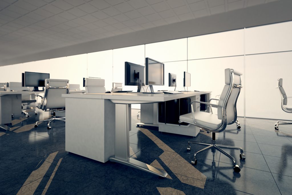 Modern office space with sleek white desks and ergonomic chairs arranged in rows. Large monitors sit atop desks, and floor-to-ceiling windows let in bright natural light, creating a clean and professional atmosphere.
