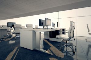Modern office space with sleek white desks and ergonomic chairs arranged in rows. Large monitors sit atop desks, and floor-to-ceiling windows let in bright natural light, creating a clean and professional atmosphere.