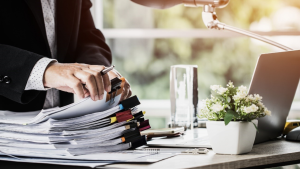 Business professional reviewing a large stack of paper documents at an office desk, highlighting records management, document organization, and the need for secure purge shredding of archived files.